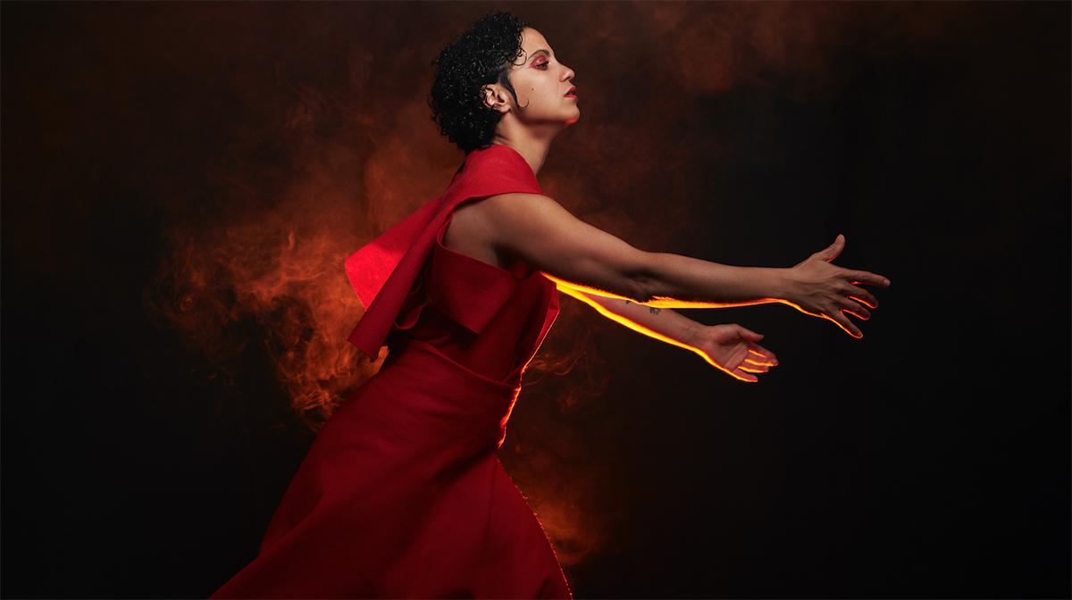 Emel Mathlouthi in profile, wearing a red dress and leaning forward with arms extended, against a dark background and red smoke.